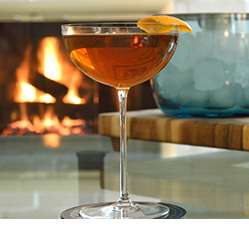 Camille Coup Chamagne Glass with a dark cocktail in it on a table in front of a fireplace