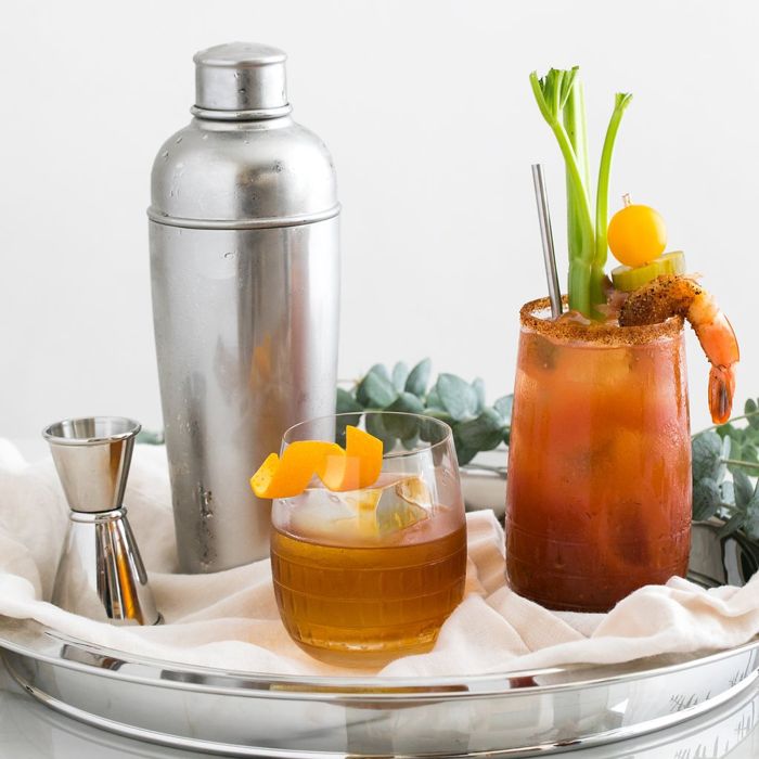 Easton Stainless Steel Cocktail Shaker next to the Ana Double Old-Fashioned Glass with an old-fashioned garnished with an orange twist on the rim, and a Bloody Mary in the Hip Highball Glass. 