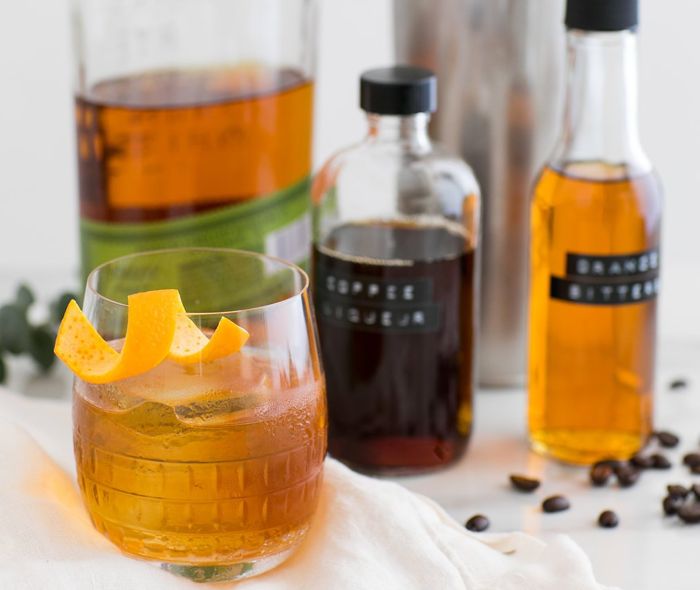 Ana Double Old-Fashioned Glass with an Old-Fashioned and an orange twist on the rim, in the background bottles with coffee liqueur and rye whiskey.