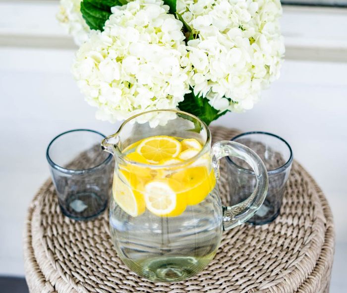 White flowers on a vase next to a set of clear glasses and a pitcher with water and lemons.