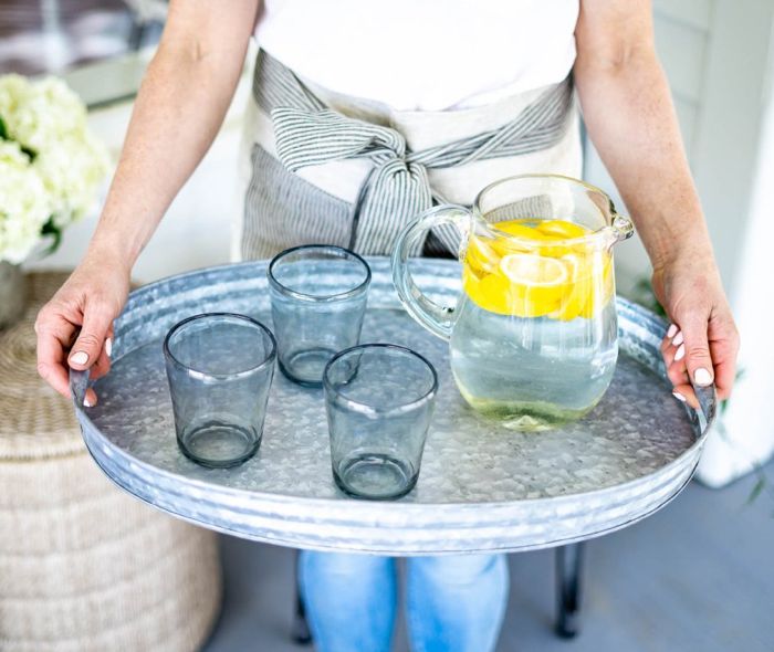 Woman holding a tray with a glass pitcher with water and cut lemons next to three clear glasses.