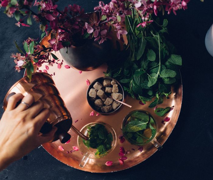 Rose gold kettle serving water to a clear mug on a rose gold tray, surrounded by flowers and herbs.