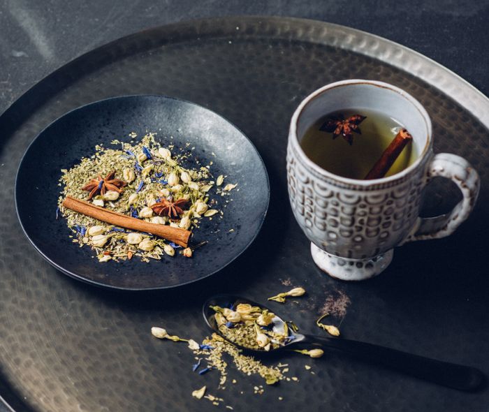  White mug with tea and cinnamon next to a black plate with the tea blend and a cinnamon stick, and a black spoon with the tea blend, over a metal serving tray