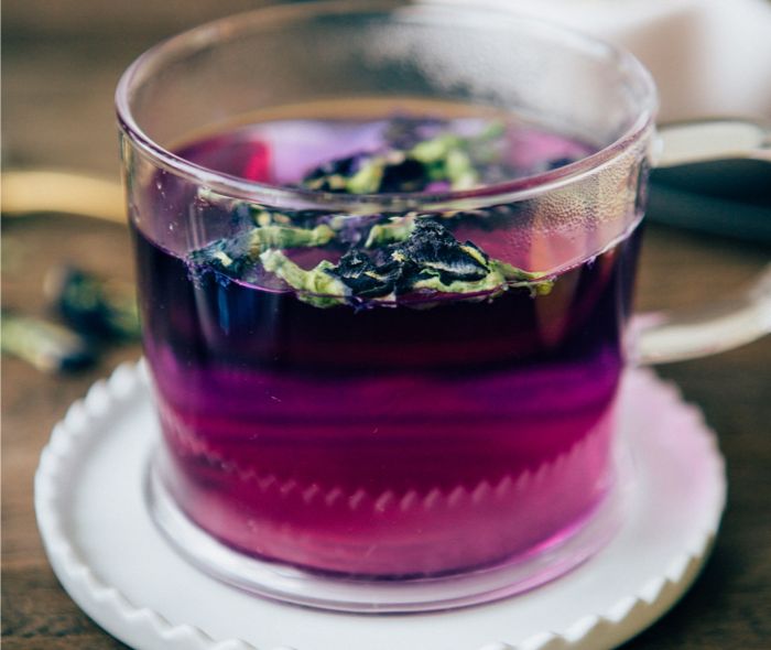 Clear mug with a purple colored Butterfly Pea Flower Tea and some herbs on top, over a white ceramic coaster.