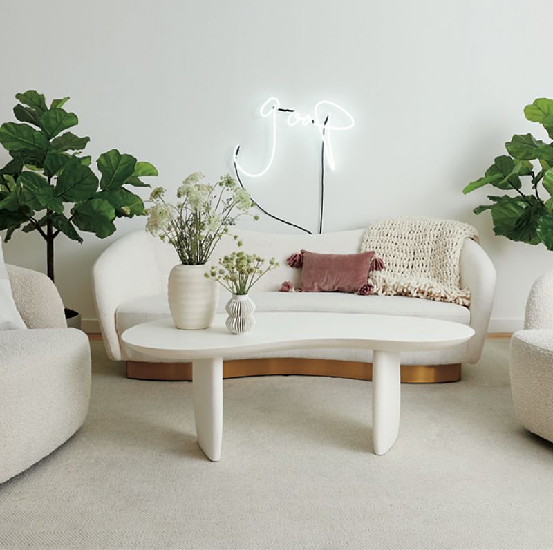 A white coffee table sits surrounded by a white couch and two white accent chairs, with a neon sign saying 'goop' hanging on the back wall