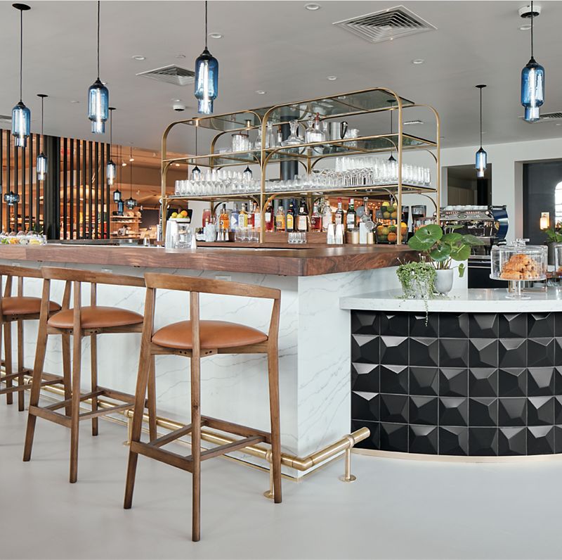 Bright bar space with liquor shelves in the background, wood bar stools  and metal pendant lights in the foreground