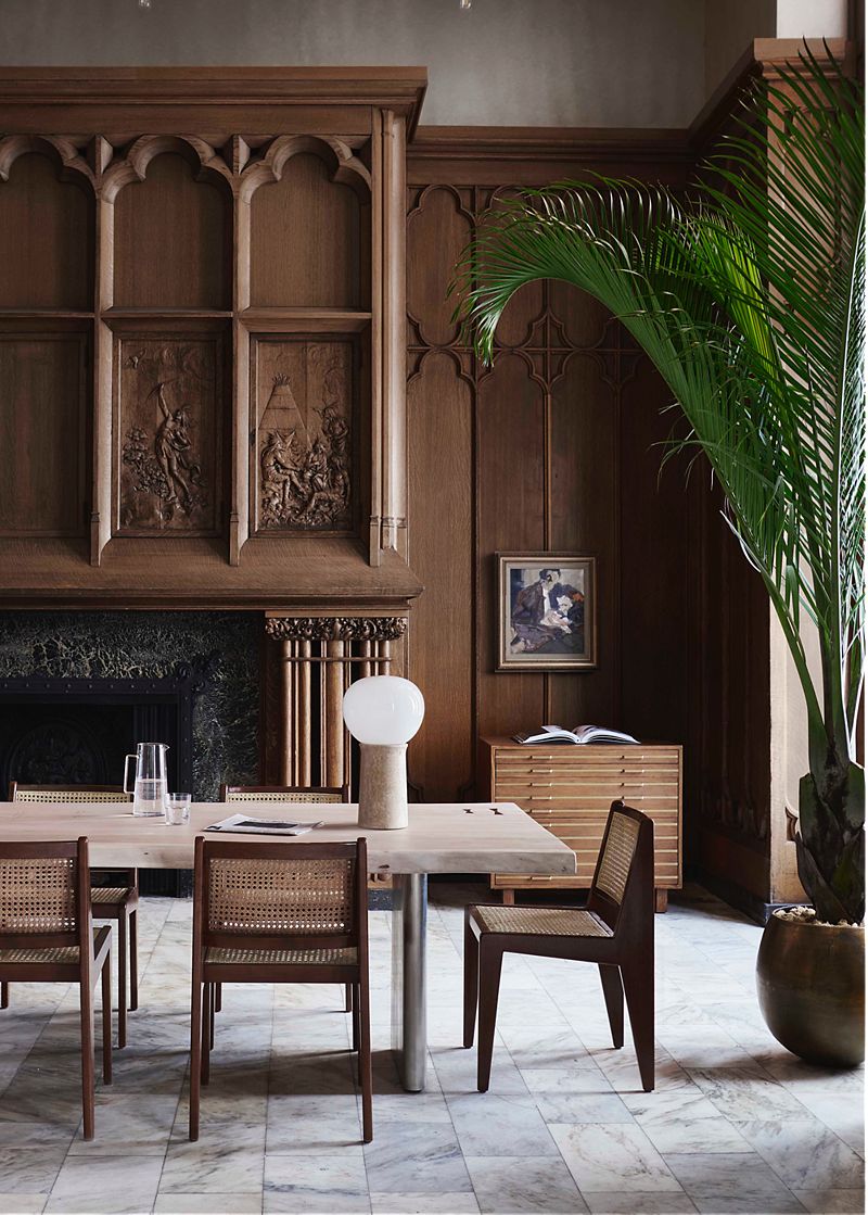 Dining space in a wood-paneled room; low cane chairs and long marble dining table in front of tall fireplace