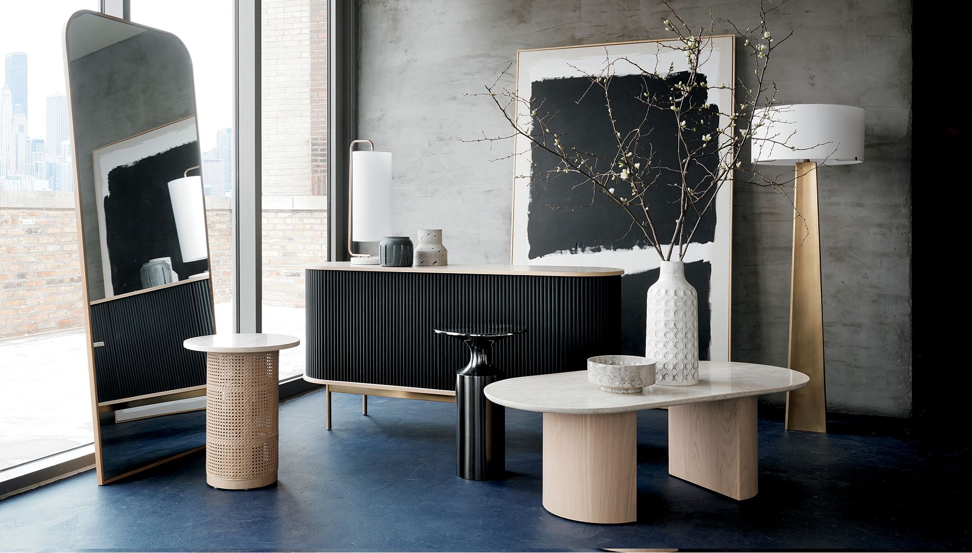 Black console table and light wood side table sit beside a standing mirror, with a large piece of art leaning against the back wall and a vase of dried branches in the foreground.