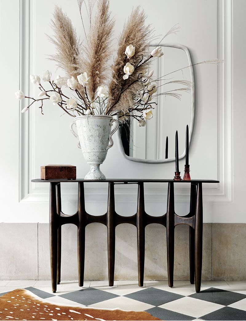 Thin brown console table holding a white vase with dried plants and two black candlesticks