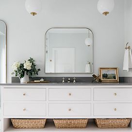 A grey bathroom counter sits on top of white drawer and brown woven baskets, with a large mirror on the center of the wall