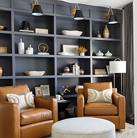 Tall black shelving sits against a wall holding various pieces of decor, and two brown leather arm chairs sit facing away from it