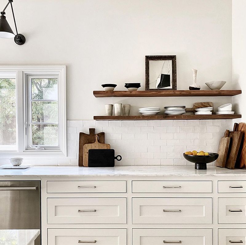 White marble kitchen counter with white drawers, and wooden shelves holding plates, bowls and cups