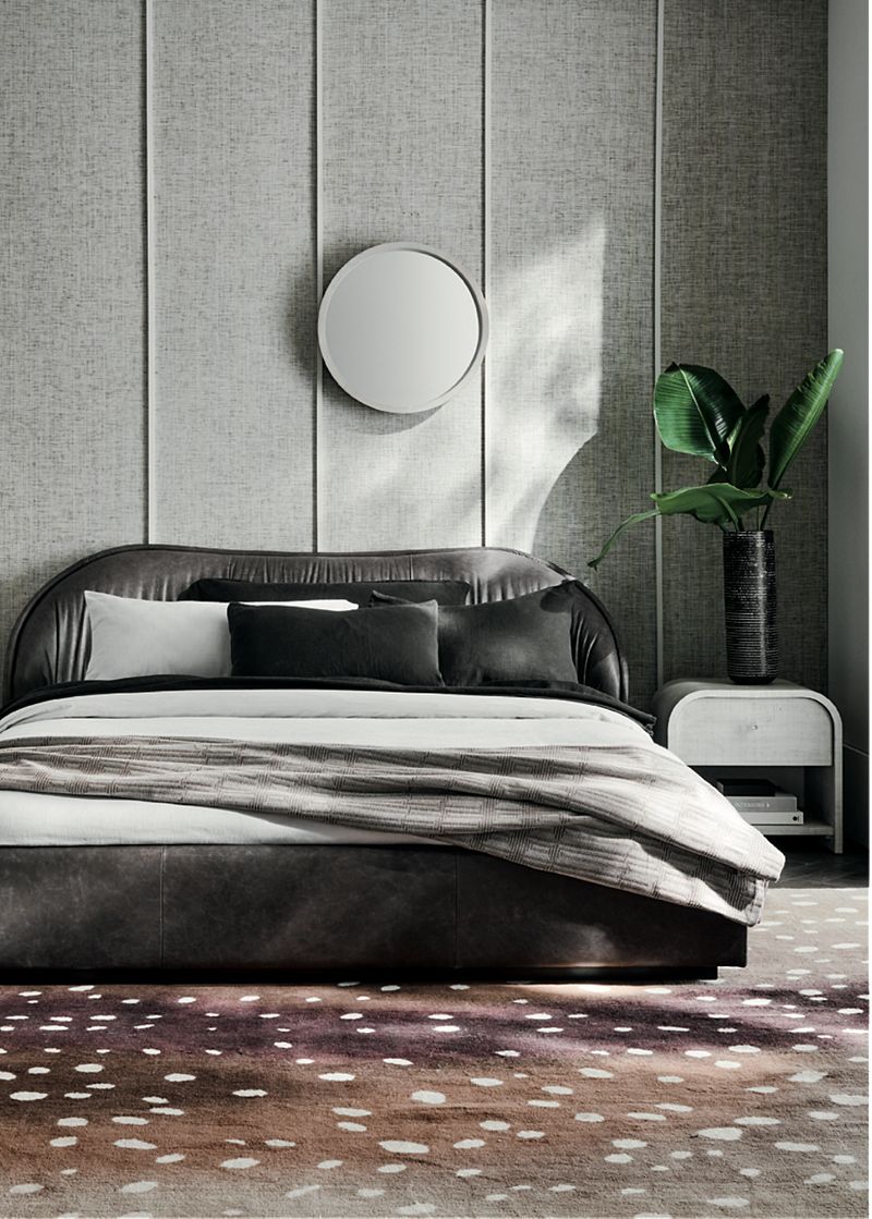 Bedroom with a low upholstered bed frame with white and grey bedding, next to which sits a white side table and plant in a black vase