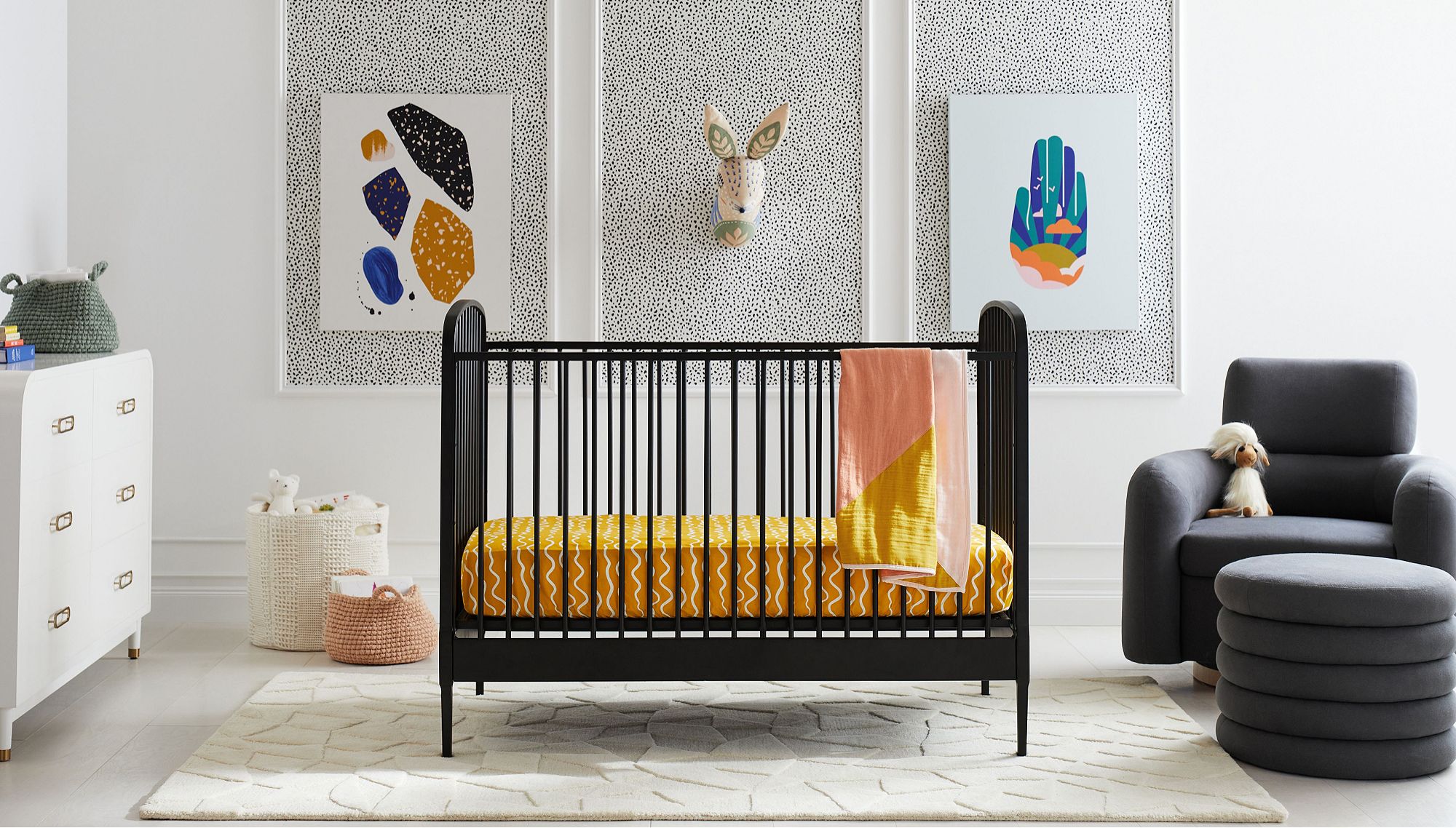 A nursery with a black crib with yellow bedding, surrounded by a white dresser, woven storage baskets and a dark grey chair