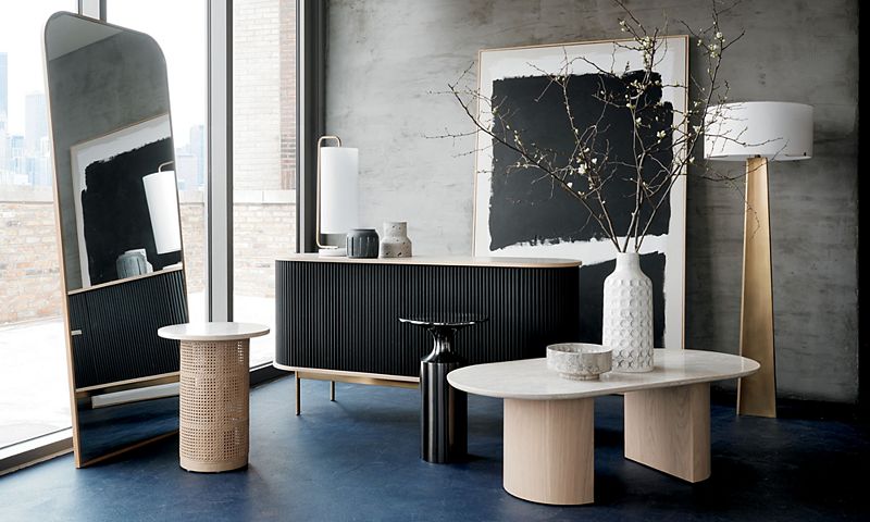 Black console table and light wood side table sit beside a standing mirror, with a large piece of art leaning against the back wall and a vase of dried branches in the foreground.