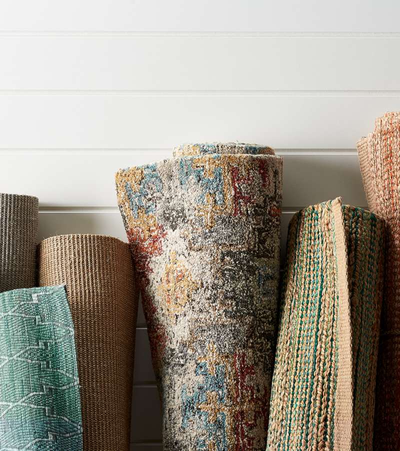 Several rolled up rugs in different sizes, patterns, colors and textures leaning up against a white paneled wall.