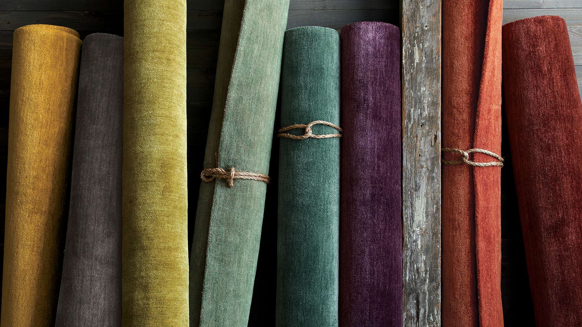 An assortment of solid-colored rugs rolled up against a wood panel wall. Shop new rugs.